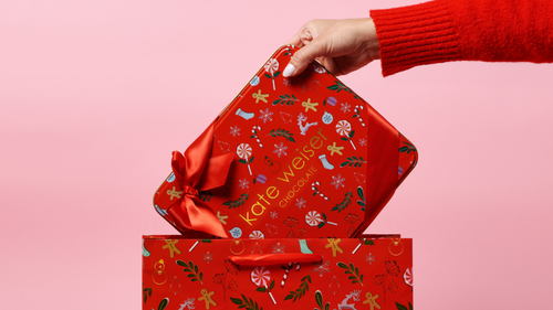 Hand opening a red gift box with a Christmas pattern against a pink background