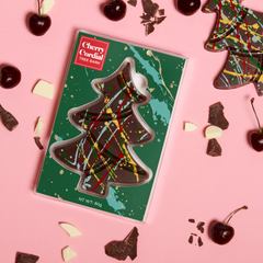 Kate Weiser's tree-shaped chocolate with colorful plaid design in green packaging, surrounded by cherries and chocolate pieces on a pink background.