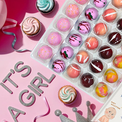 An array of colorful chocolate bonbons in various flavors, arranged on a tray with a pink background, celebrating a baby shower with 'IT'S A GIRL' text visible.